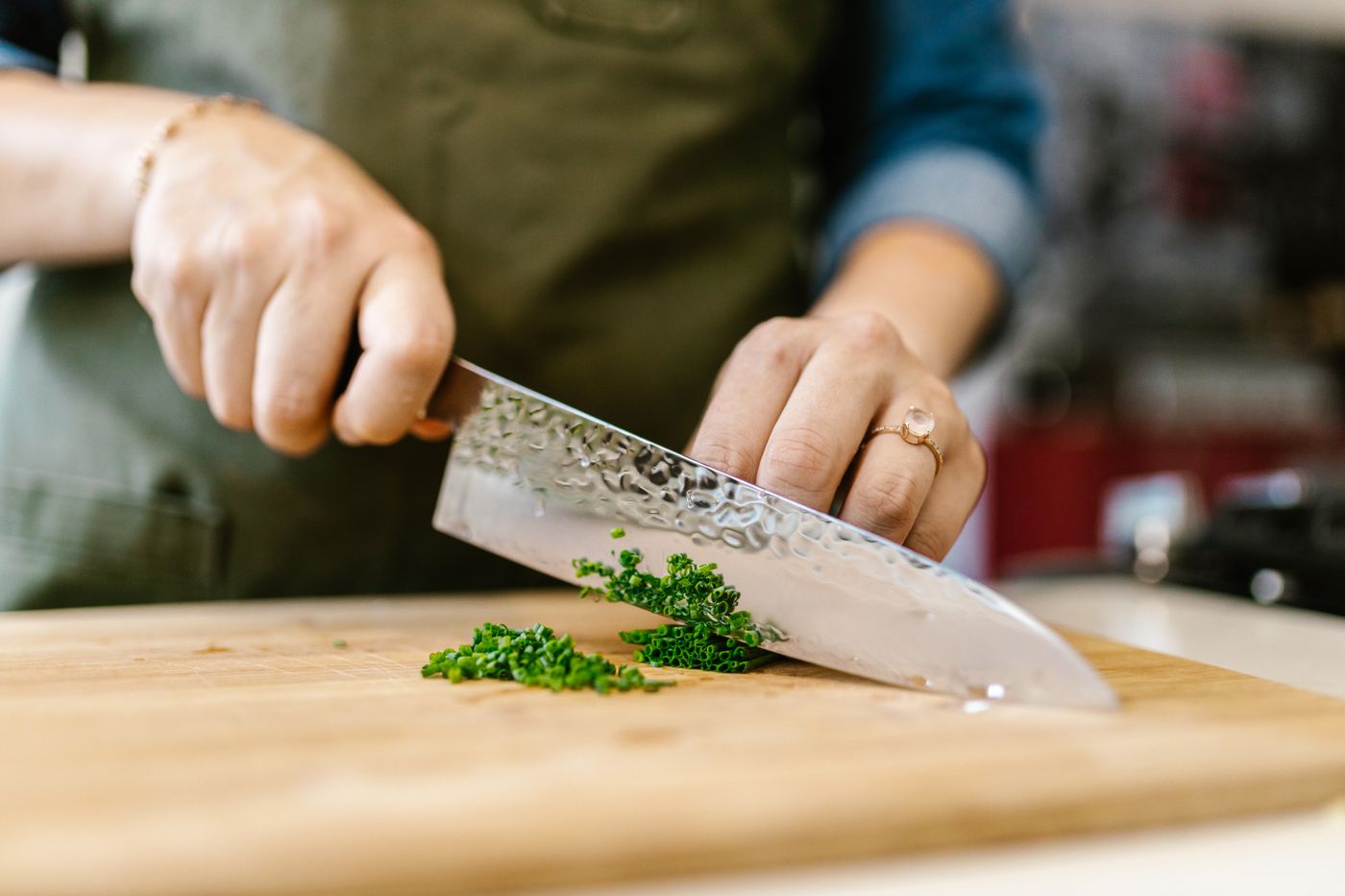 Hands finely chopping fresh chives with a chef’s knife on a wooden prep board.